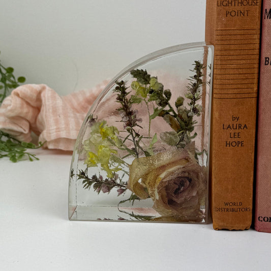 Glass bookend with preserved flowers next to a stack of books on a white surface.