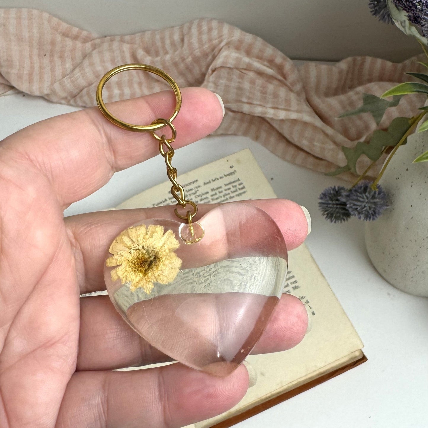 Hand holding a keychain with a clear heart-shaped pendant and dried flower, on a white surface with a pink textured fabric and decorative elements in the background.